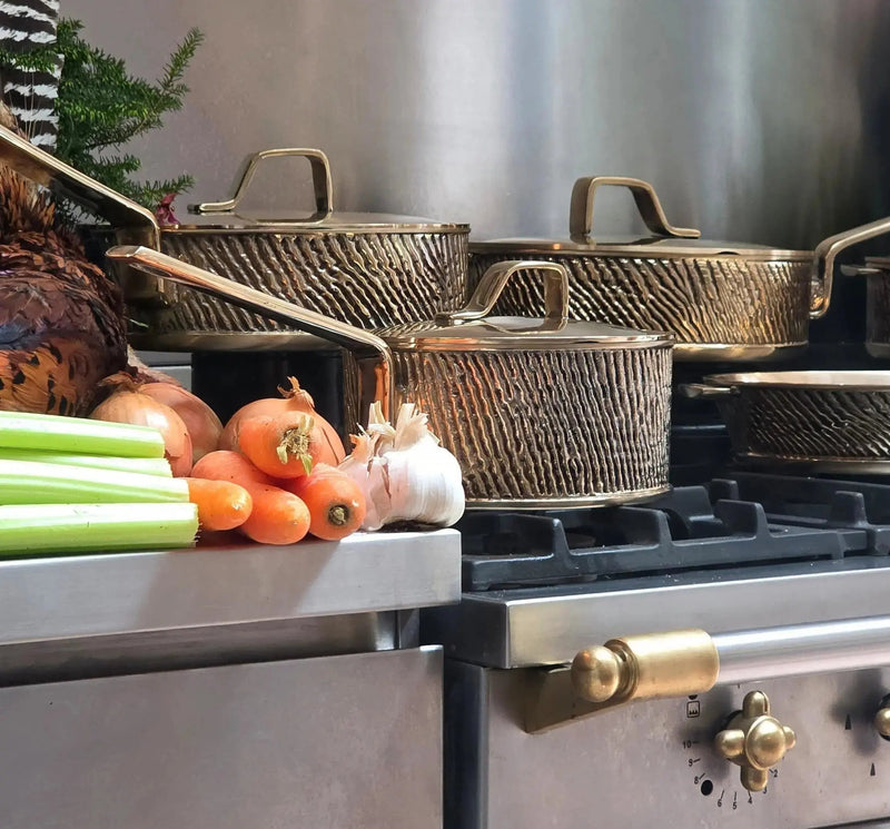 "Eaglador heavy gauge Kansa bronze cookware collection seen on a stove top with vegetables on the side - Luxury Cookware UK".